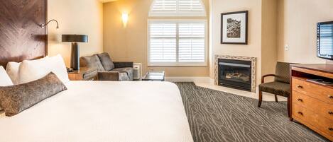A hotel room with a large bed, a desk, a chair, a fireplace, and a window with blinds.