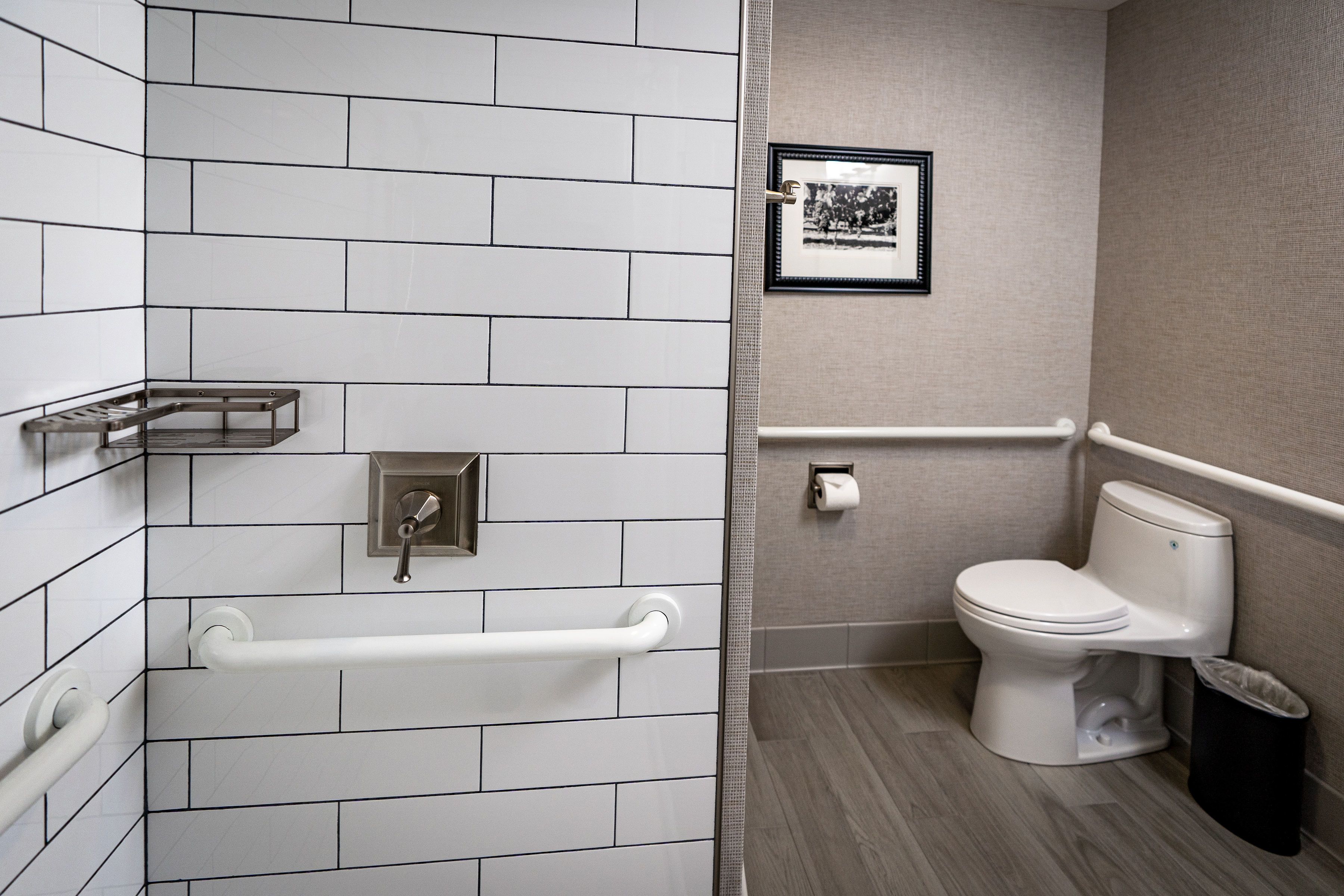 A bathroom with white tiled walls, a white toilet, a handrail, a trash bin, and a framed picture on the wall.