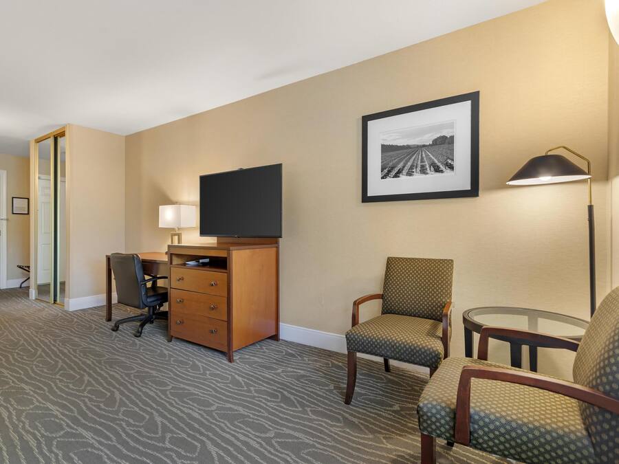 A hotel room with a flat-screen TV, a desk, two chairs, a small table, a lamp, and a framed picture on the wall.