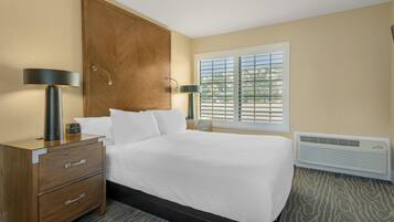 Quarto de hotel com uma cama, uma mesa de cabeceira de madeira com gavetas, uma luminária, uma janela com persianas e um ar condicionado.