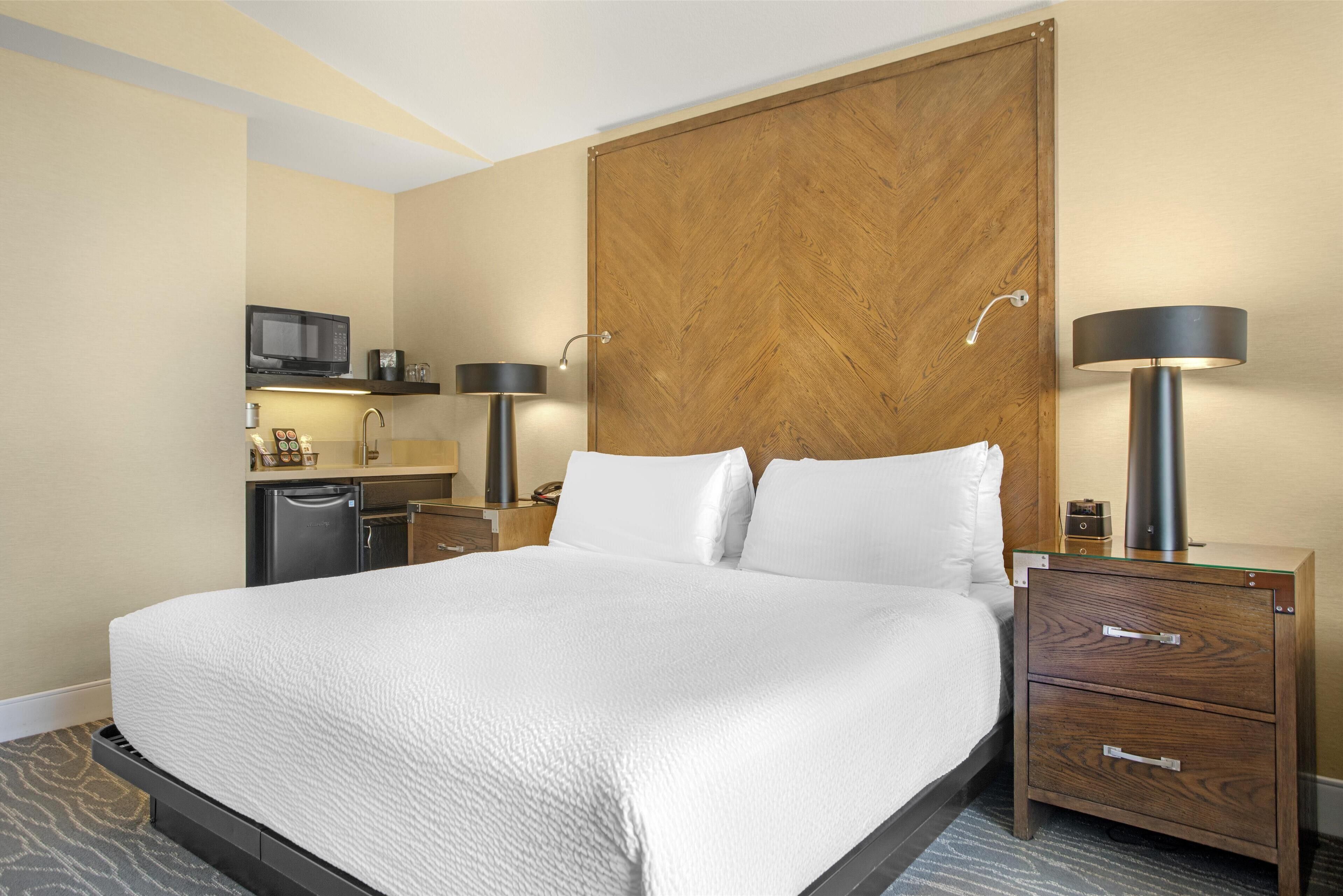 A hotel room with a large bed, a wooden wardrobe, a desk with a lamp, a microwave, and a small kitchenette.