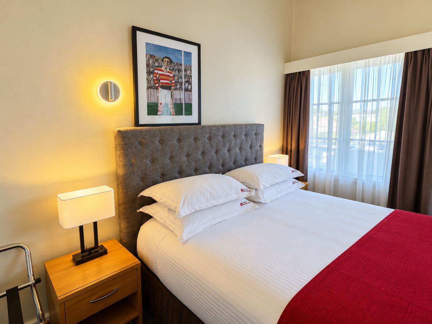 A hotel room with a bed, a nightstand, a framed picture on the wall, and a window with curtains.