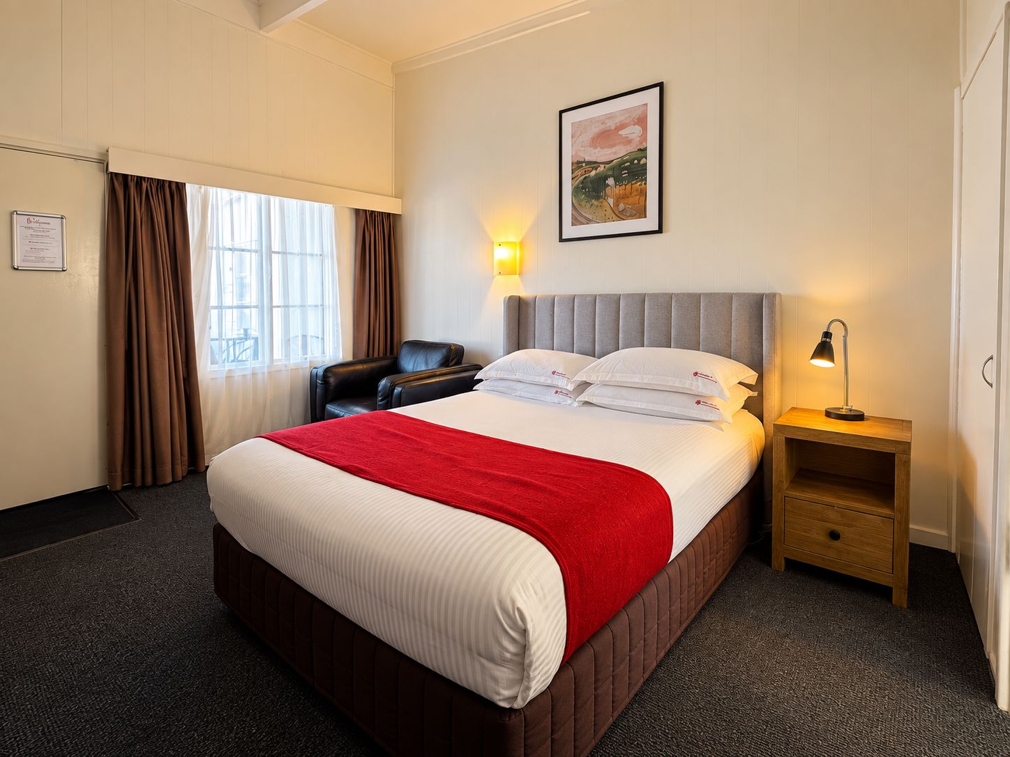A hotel room with a large bed, a bedside table with a lamp, a chair, a window with curtains, and a framed picture on the wall.