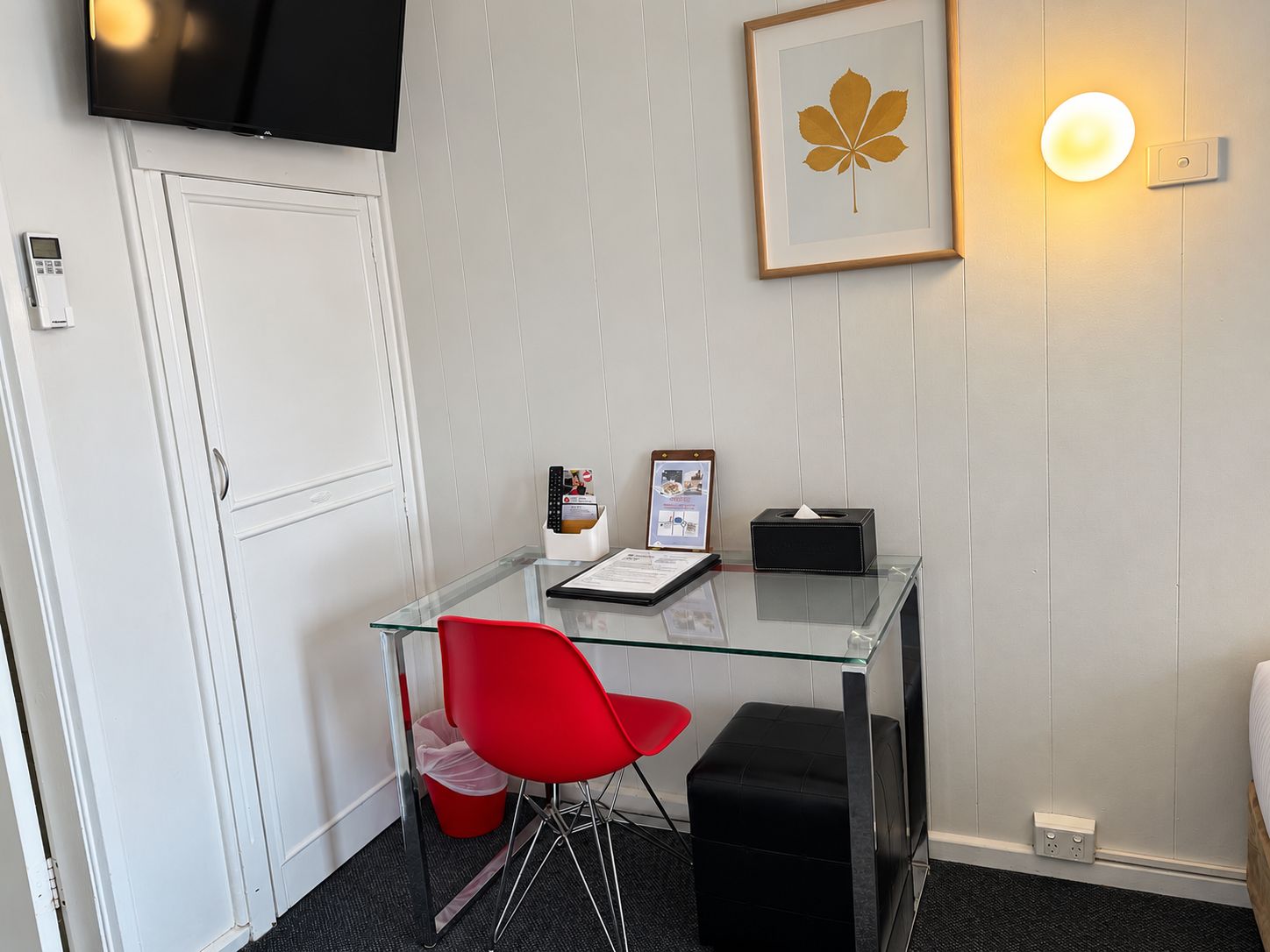 A hotel room with a glass desk, a red chair, a black ottoman, a framed leaf artwork, a wall-mounted light, and a television.