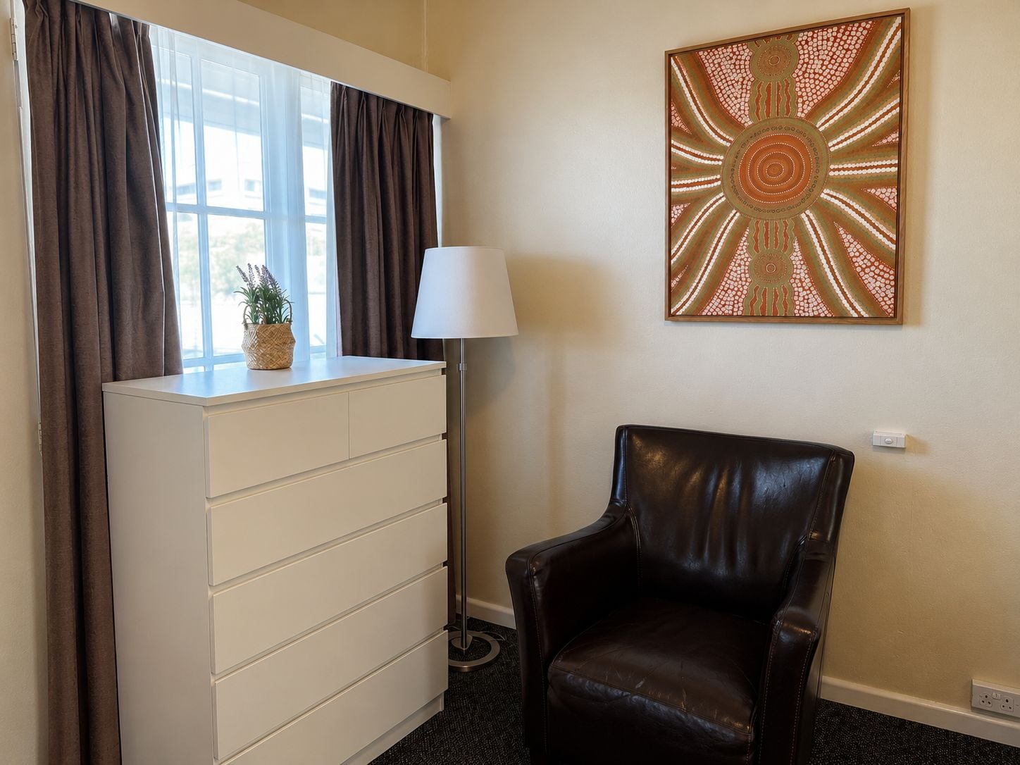 A room with a white dresser, a brown leather chair, a floor lamp, and a wall art piece.