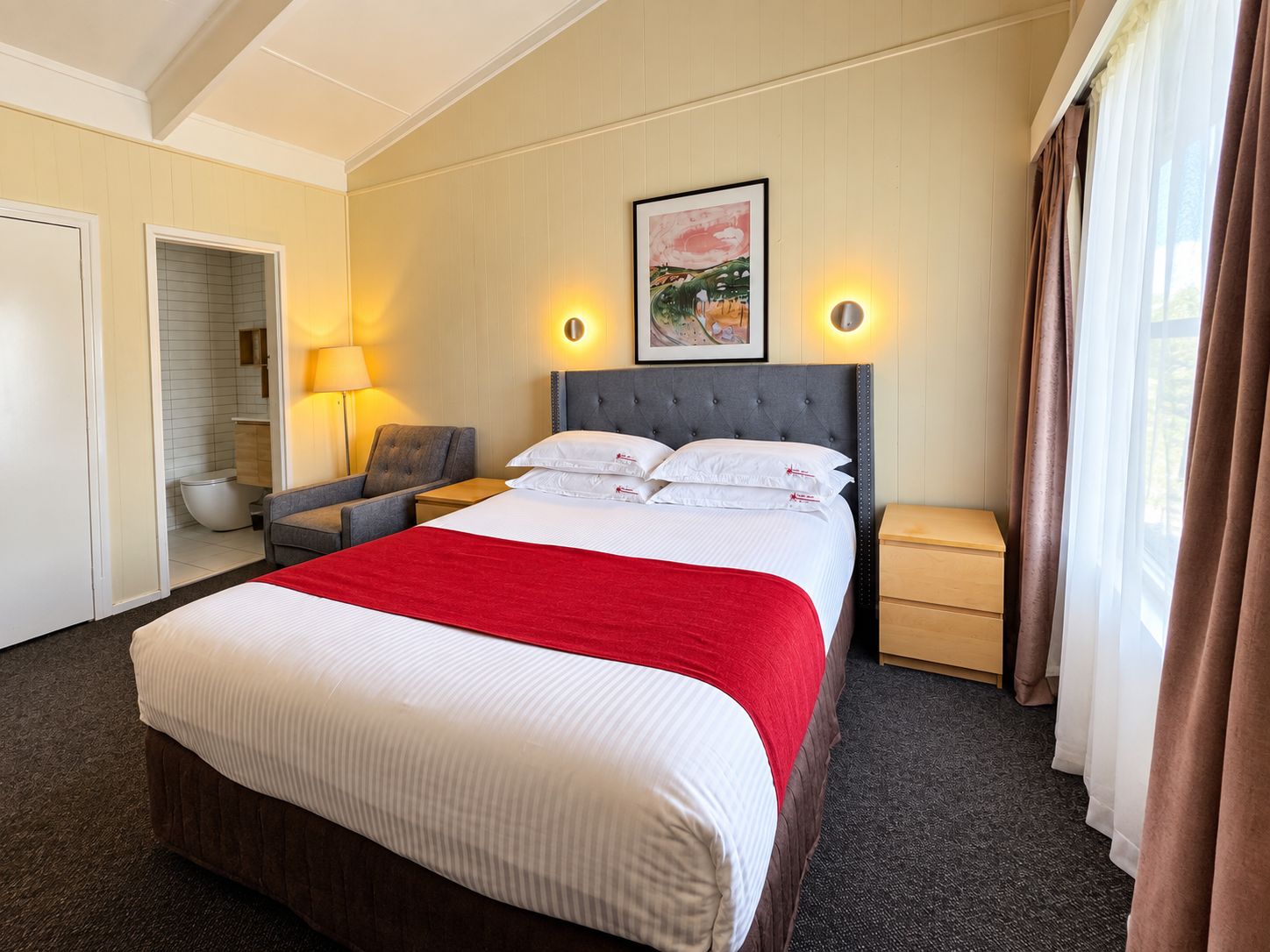 Habitación de hotel con una cama grande, una silla, una mesita de noche, una lámpara y un cuadro en la pared.