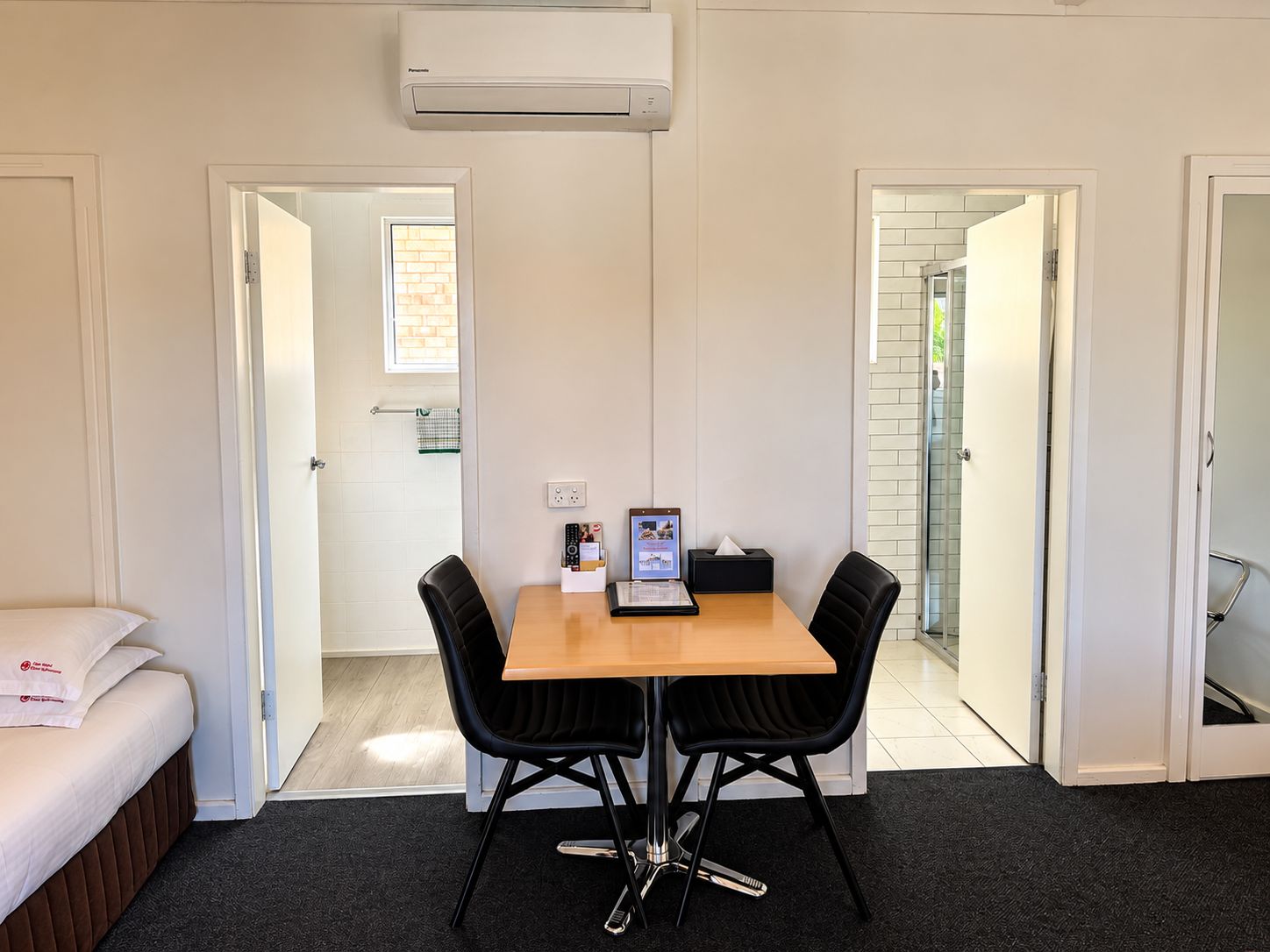 A hotel room with a bed, a dining table with chairs, an air conditioning unit, and a view of a brick wall through an open door.