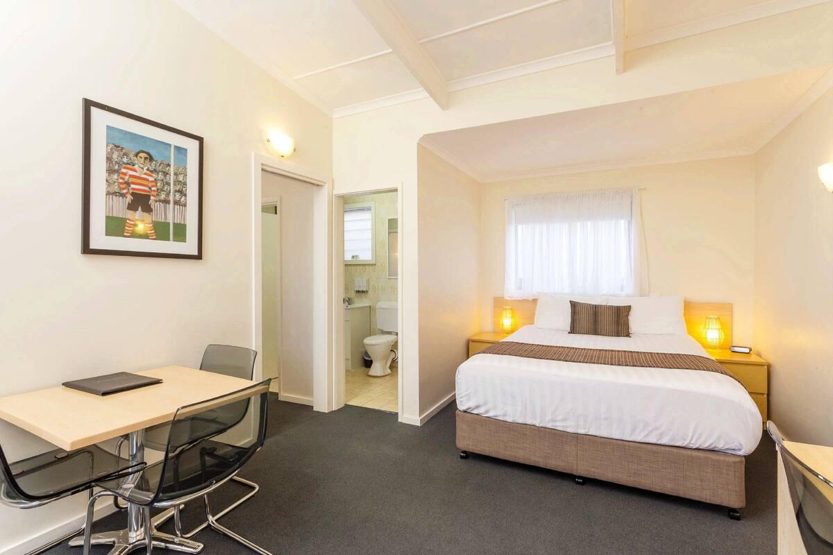 A hotel room with a bed, bedside lamps, a desk with a chair, a framed picture on the wall, and a bathroom visible through an open door.