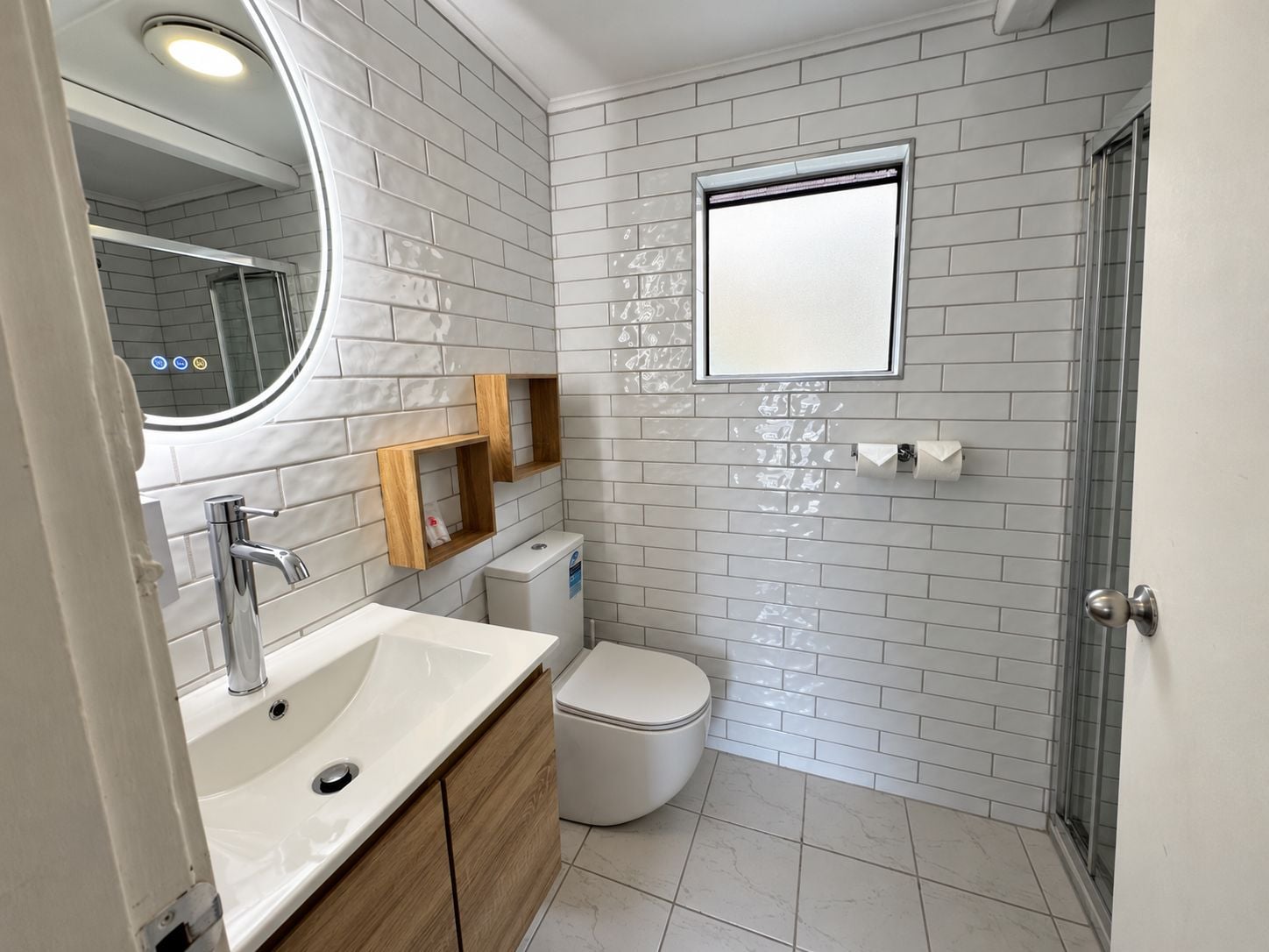 Une salle de bain moderne avec un lavabo, un miroir et des toilettes blancs, agrémentée d’un mur recouvert de carreaux et d’une petite fenêtre.