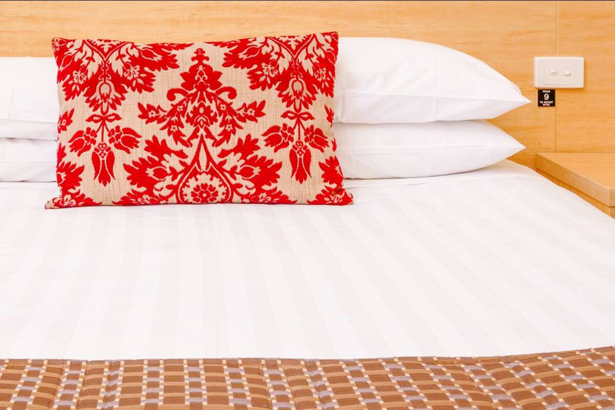 A neatly made bed with white linens, a red and white patterned pillow, and a brown woven throw at the foot of the bed. There are two bedside tables with a clock and a phone on each.