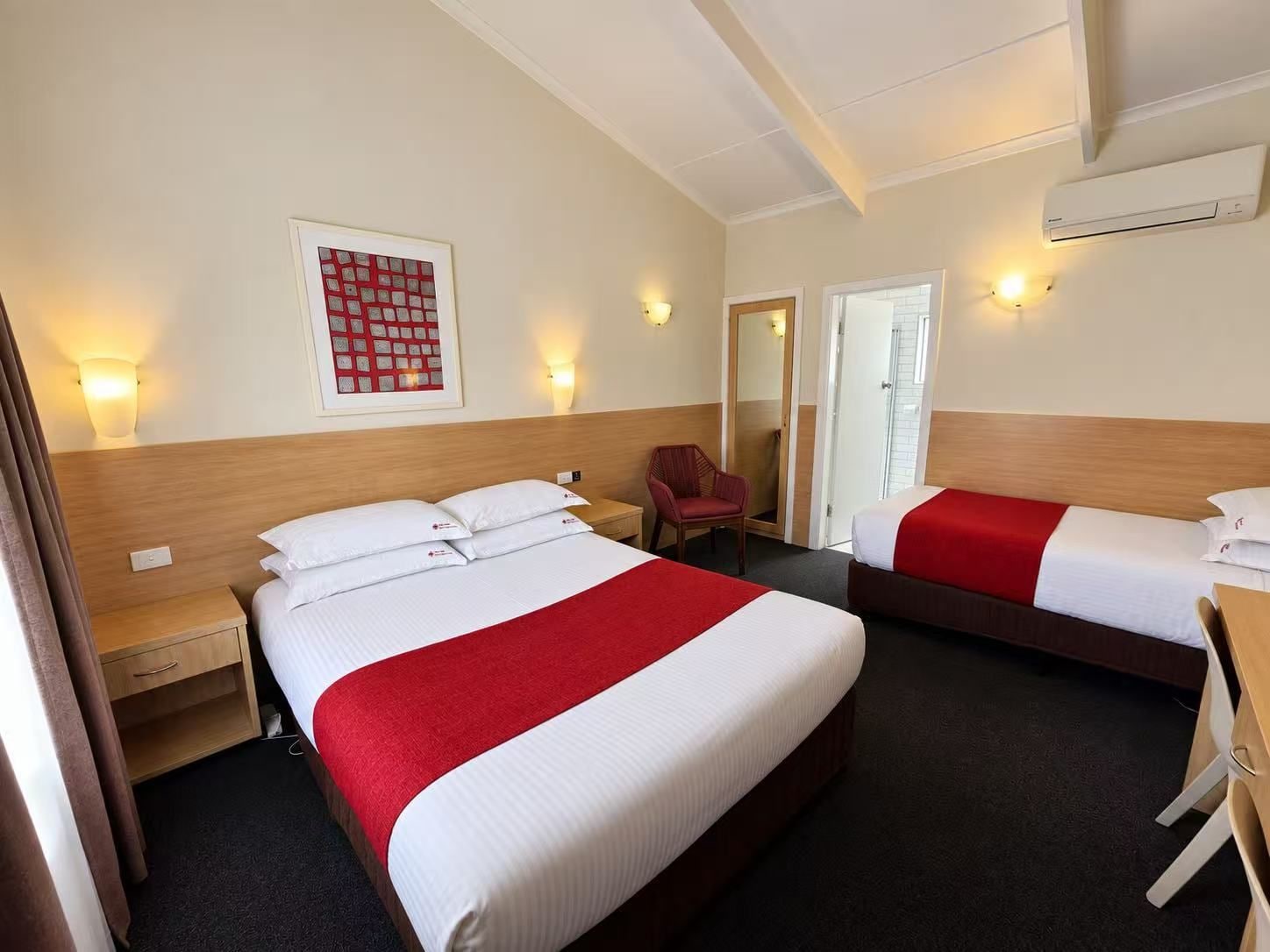 A hotel room with two beds, a chair, a desk, and a wall-mounted air conditioner.