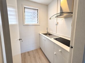 A compact kitchen with white tiles, a window, a stainless steel sink, and a black induction cooktop. - Club Motel (Wagga Wagga)
