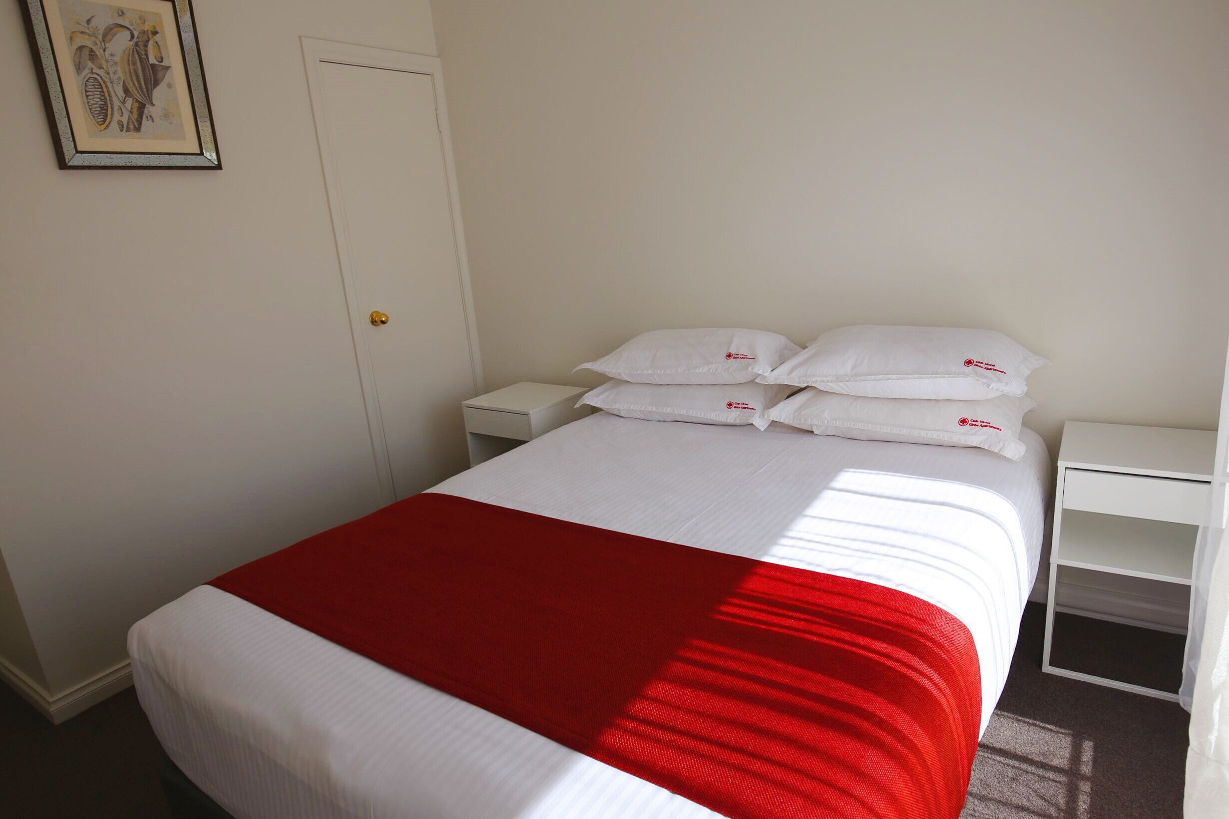 A neatly made bed with white bedding and a red accent, a white nightstand with a lamp, and a white wall with a framed picture.
