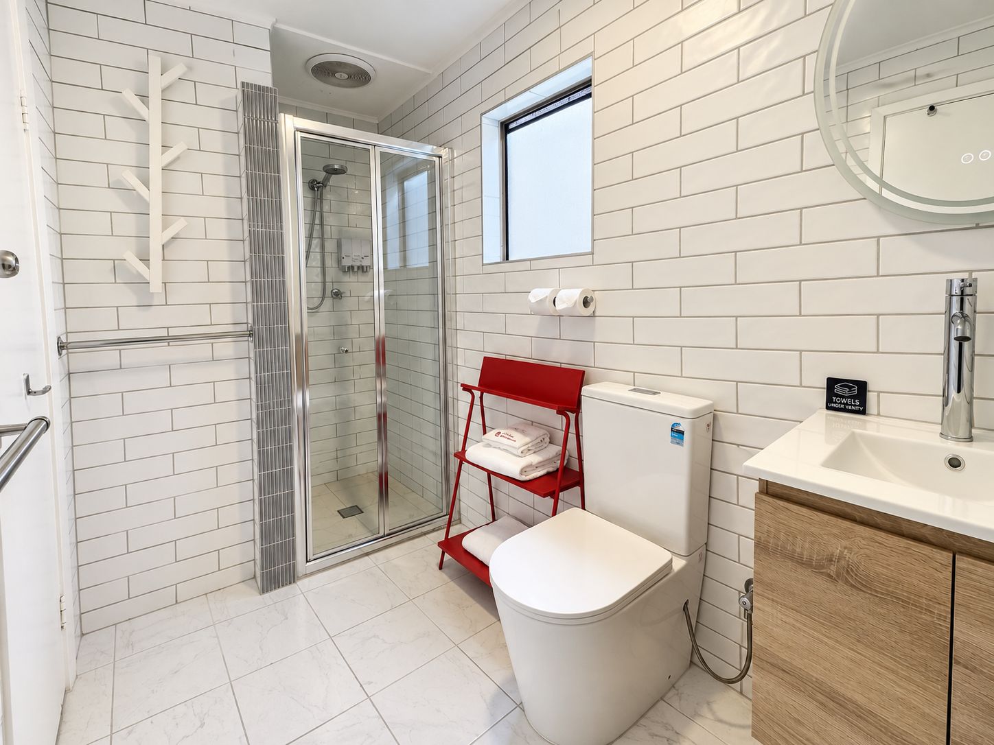 Et moderne badeværelse med en brusekabine i glas, toilet, håndvask og en rød stol.