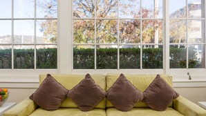 A room with a yellow sofa and brown cushions, large windows with a view of trees and a hedge. - Club Motel (Wagga Wagga)