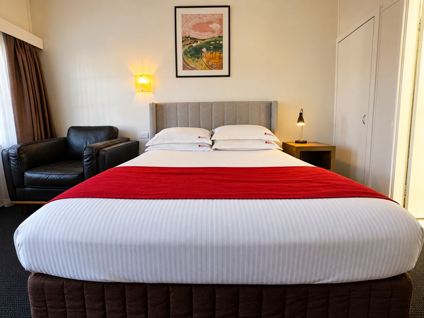 A hotel room with a bed, a chair, a nightstand, a lamp, and a painting on the wall.