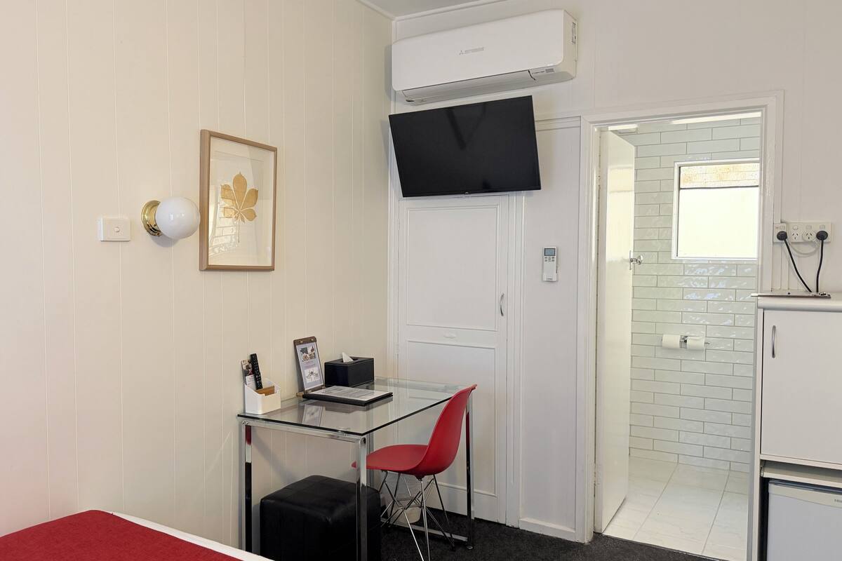 A compact room with a glass desk, a red chair, a framed picture, a black TV, and a white air conditioning unit.