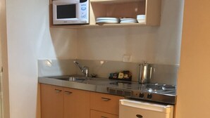 Eine kompakte Küche mit Mikrowelle, Herd und Spüle. Ein kleiner Kühlschrank und ein Regal über der Arbeitsfläche sind vorhanden. Auf dem Tisch steht eine Orchidee im Topf.