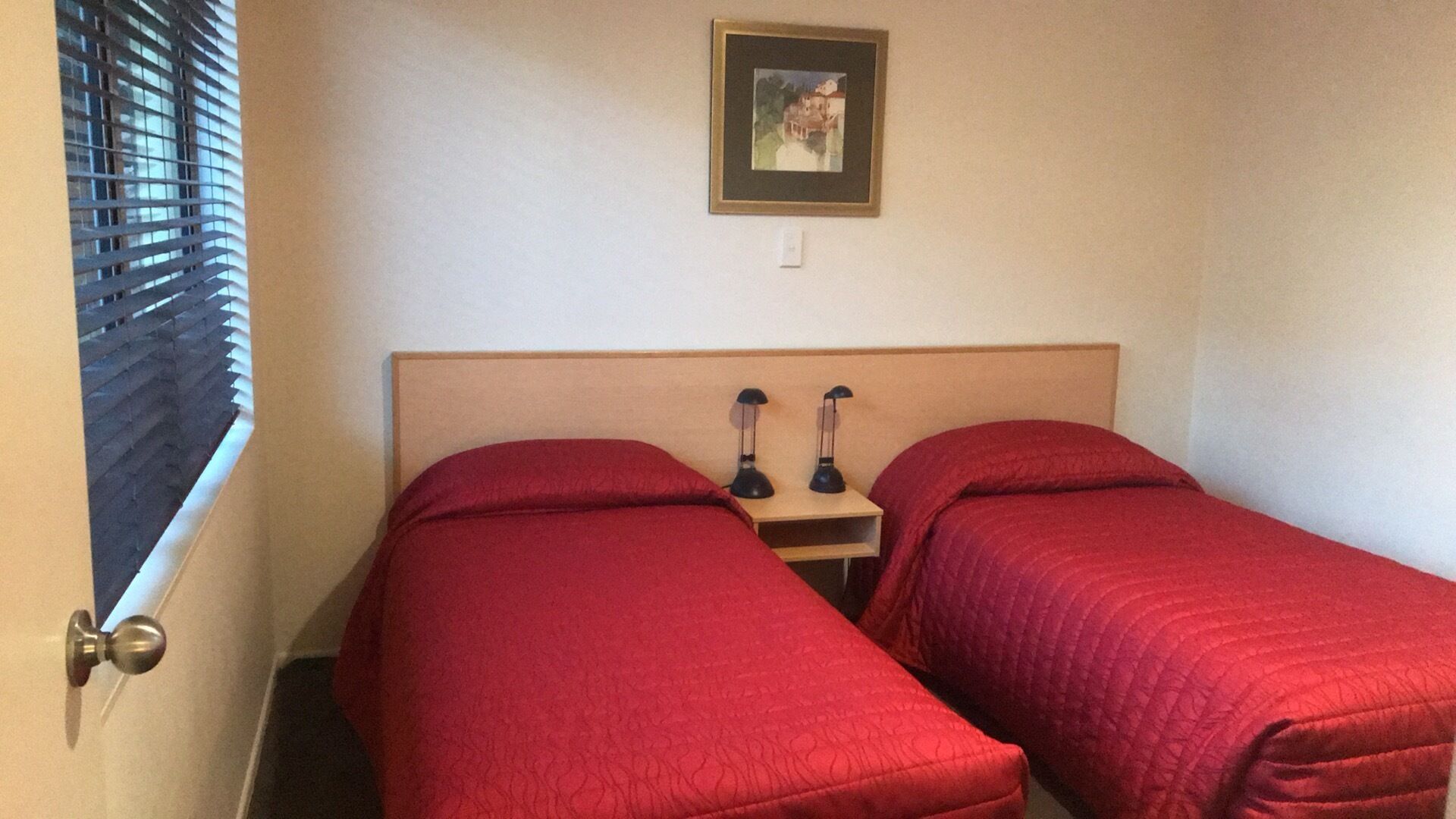 A room with two single beds, each with red bedding, a wooden headboard, and a small bedside table with two black lamps.