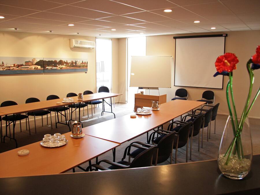 A conference room with long tables, chairs, a projector screen, and a large window with curtains.