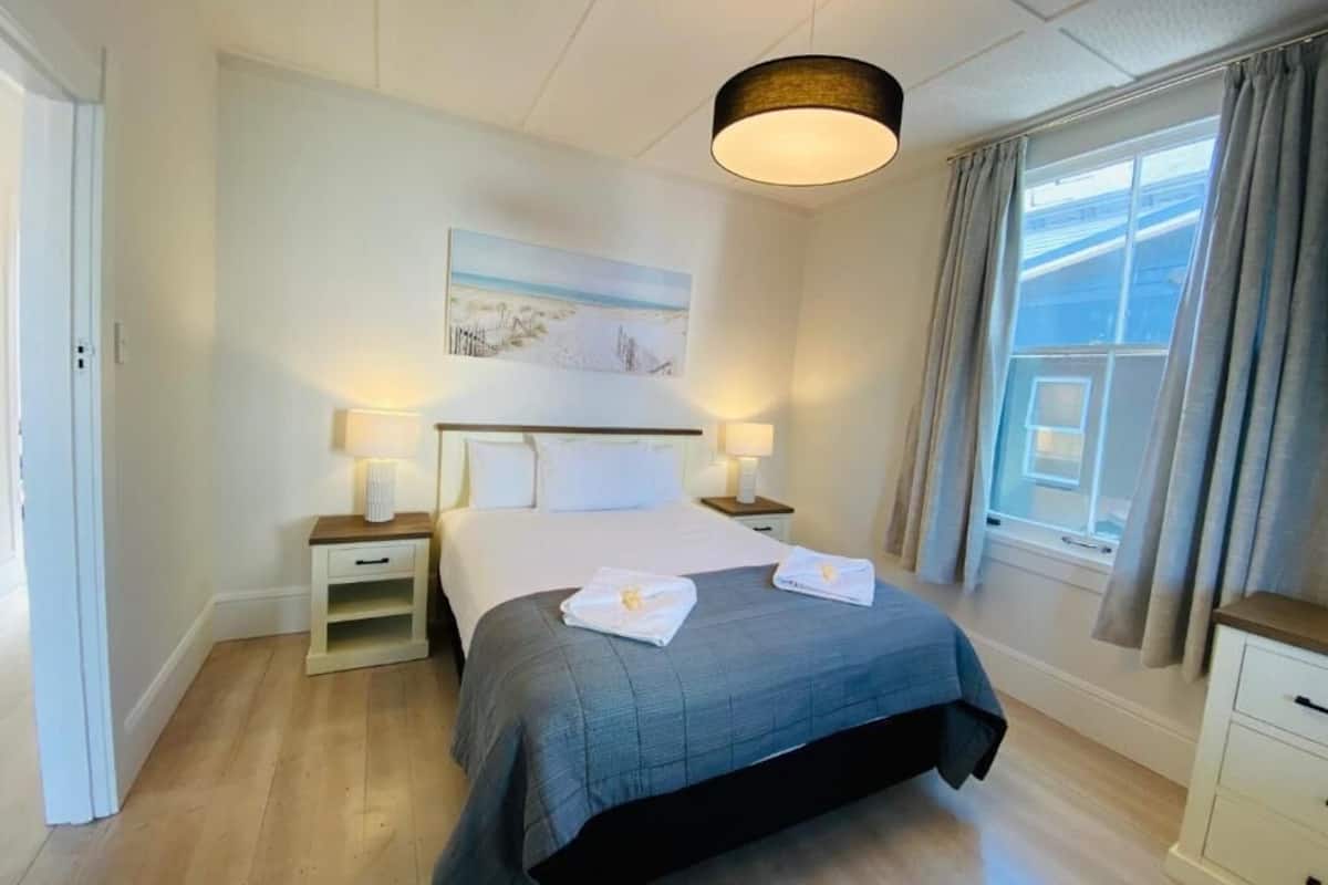 Una habitación de hotel con cama, mesitas de noche, un cuadro en la pared, una ventana con cortinas y una lámpara colgante.