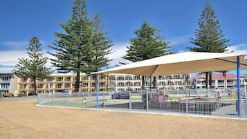 A fenced sports field with a pavilion, surrounded by tall trees and a building in the background.
