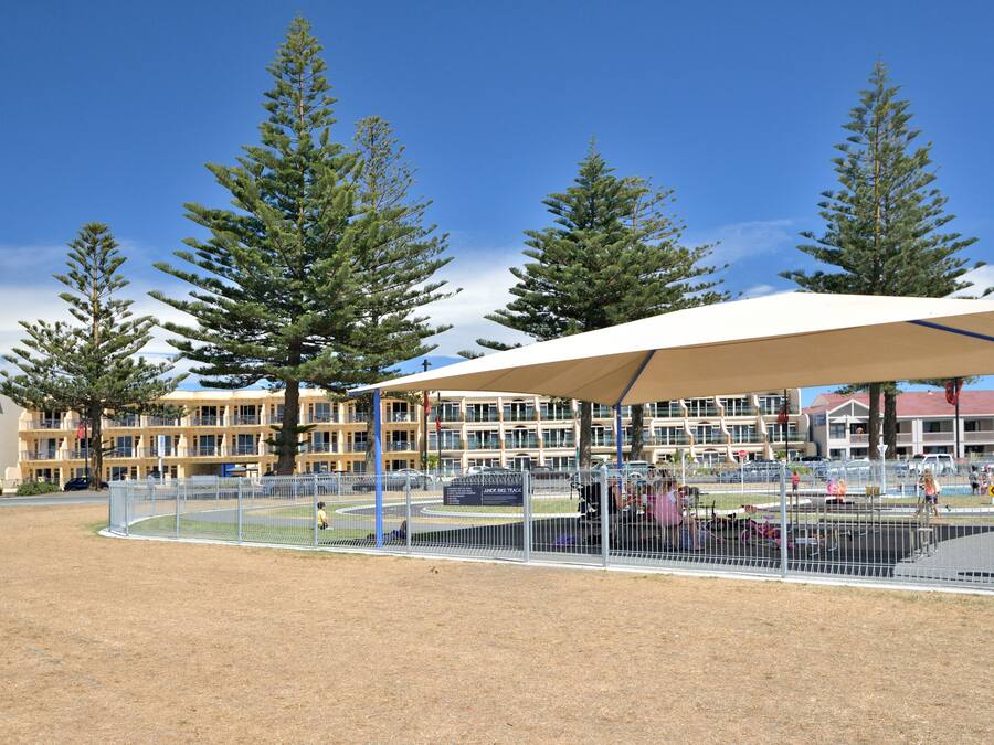 A fenced sports field with a pavilion, surrounded by tall trees and a building in the background.