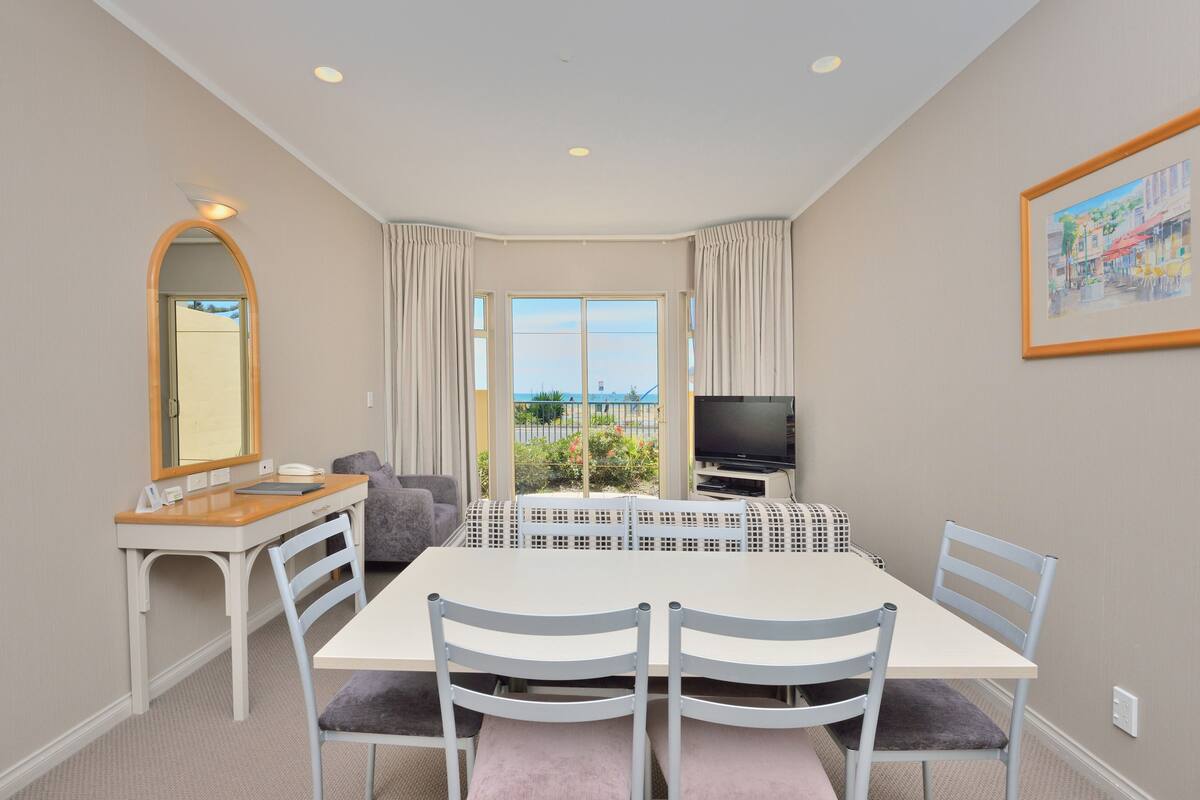 Una habitación con una mesa de comedor, sillas, un televisor, un escritorio con espejo y una ventana con cortinas.