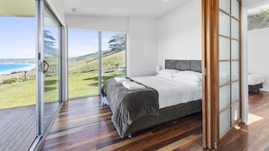 A bedroom with a large bed, a sliding glass door leading to a balcony with a view of the sea, and a wooden floor. - Seafarers Getaway (Apollo Bay)
