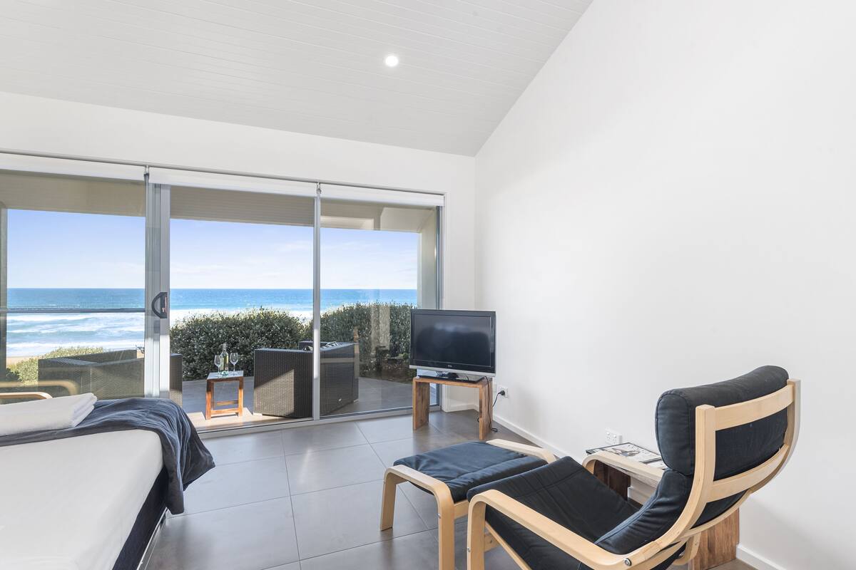 A modern room with a bed, a TV on a stand, and a chair. There's a large sliding glass door leading to a balcony with a view of the ocean.