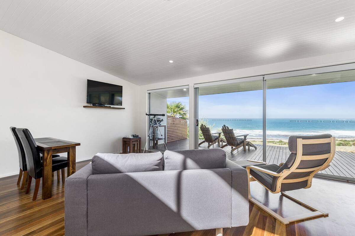 A modern living room with a gray sofa, wooden dining table, and chairs, a TV mounted on the wall, and a large sliding glass door leading to a balcony with a view of the ocean.