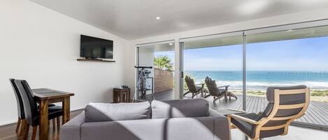 A modern living room with a gray sofa, wooden dining table, and chairs, a TV mounted on the wall, and a large sliding glass door leading to a balcony with a view of the ocean.