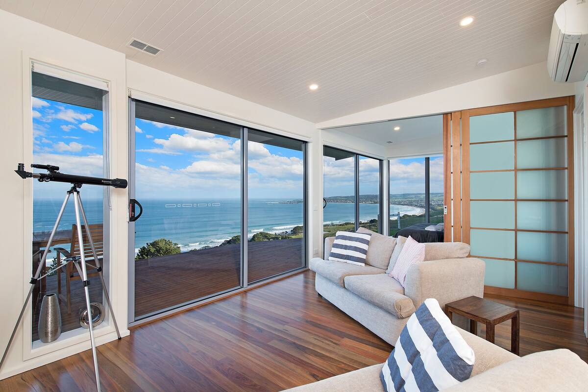 A modern living room with a large sliding glass door leading to a balcony overlooking the ocean.