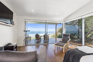 A modern room with a large sliding glass door leading to a deck with a view of the ocean. There is a sofa, a chair, and a small table with chess pieces.