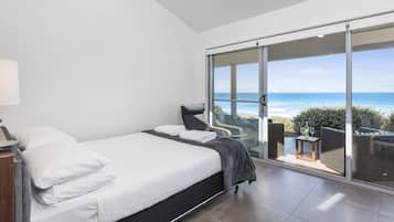 A modern bedroom with a large bed, a chair, and a view of the ocean through a sliding glass door.