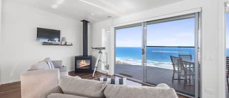 A modern living room with a sofa, a fireplace, a TV, and a large sliding glass door leading to a balcony with a view of the ocean.