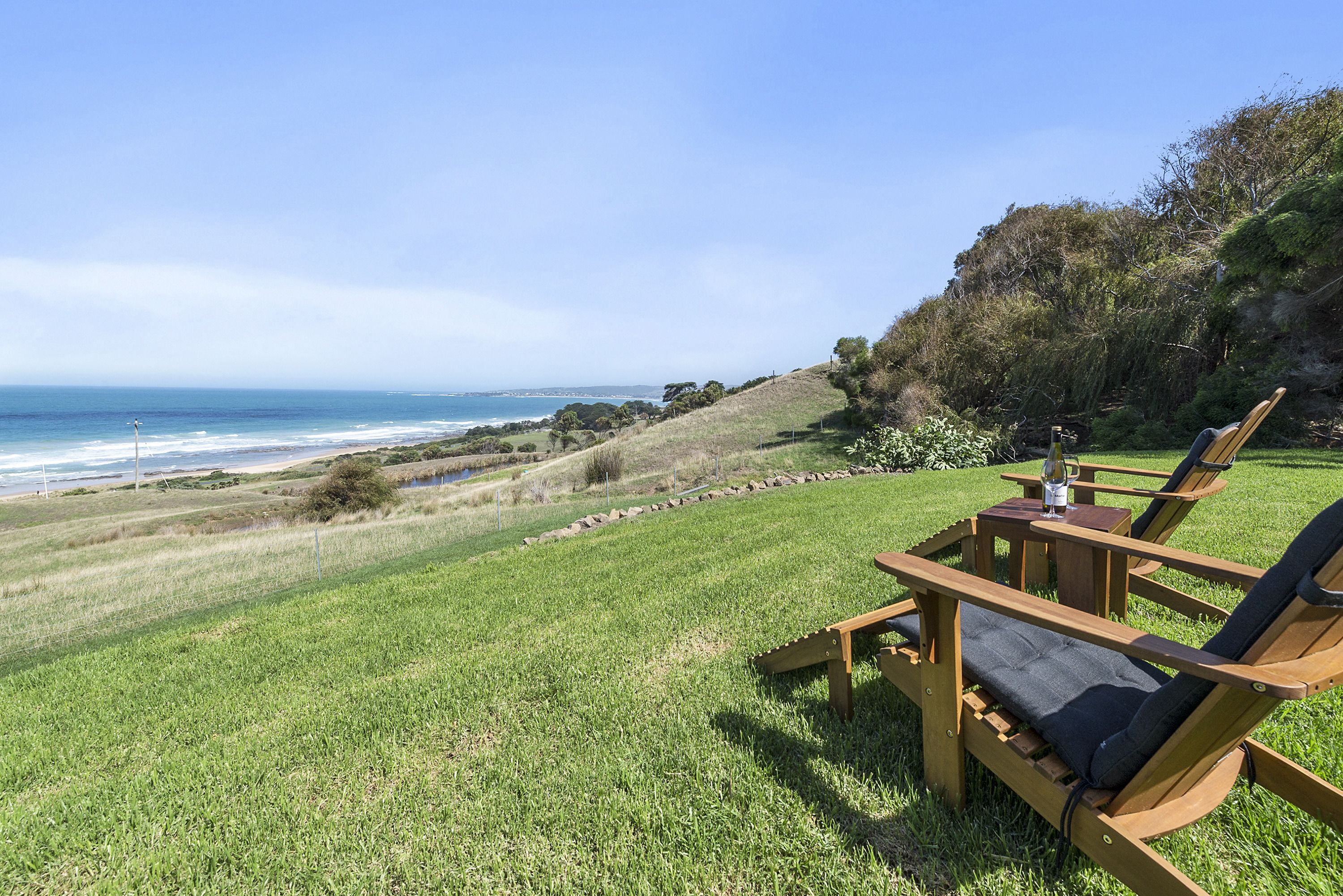 Zona verde con una silla y mesa de madera, con vista a una playa y al océano.