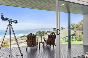 A telescope on a tripod, two Adirondack chairs, a small table with a bottle and glass, and a view of the ocean and hills.