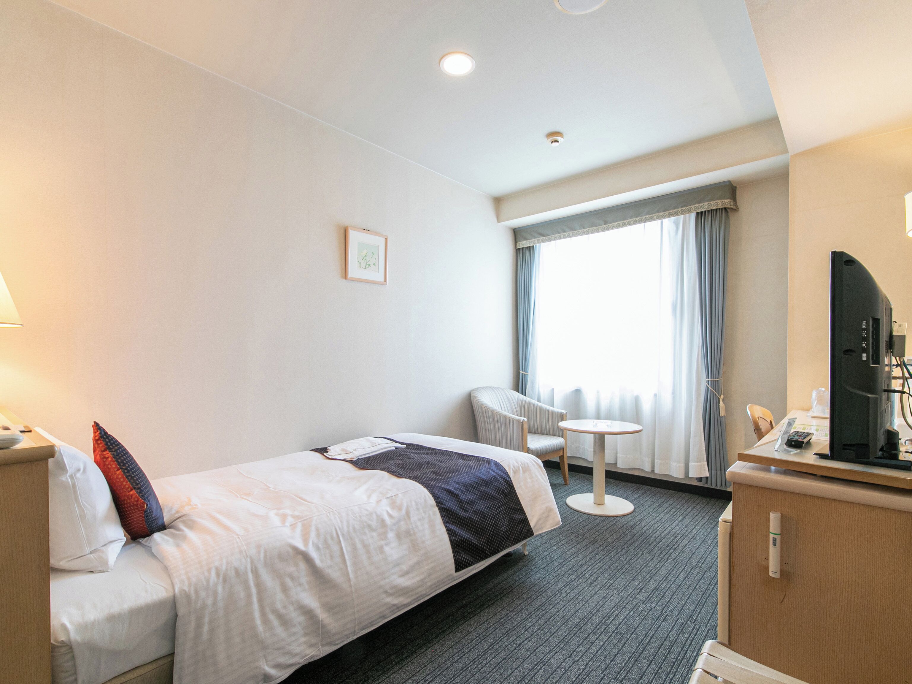 A hotel room with a bed, a small round table, a chair, a television, and a window with curtains.