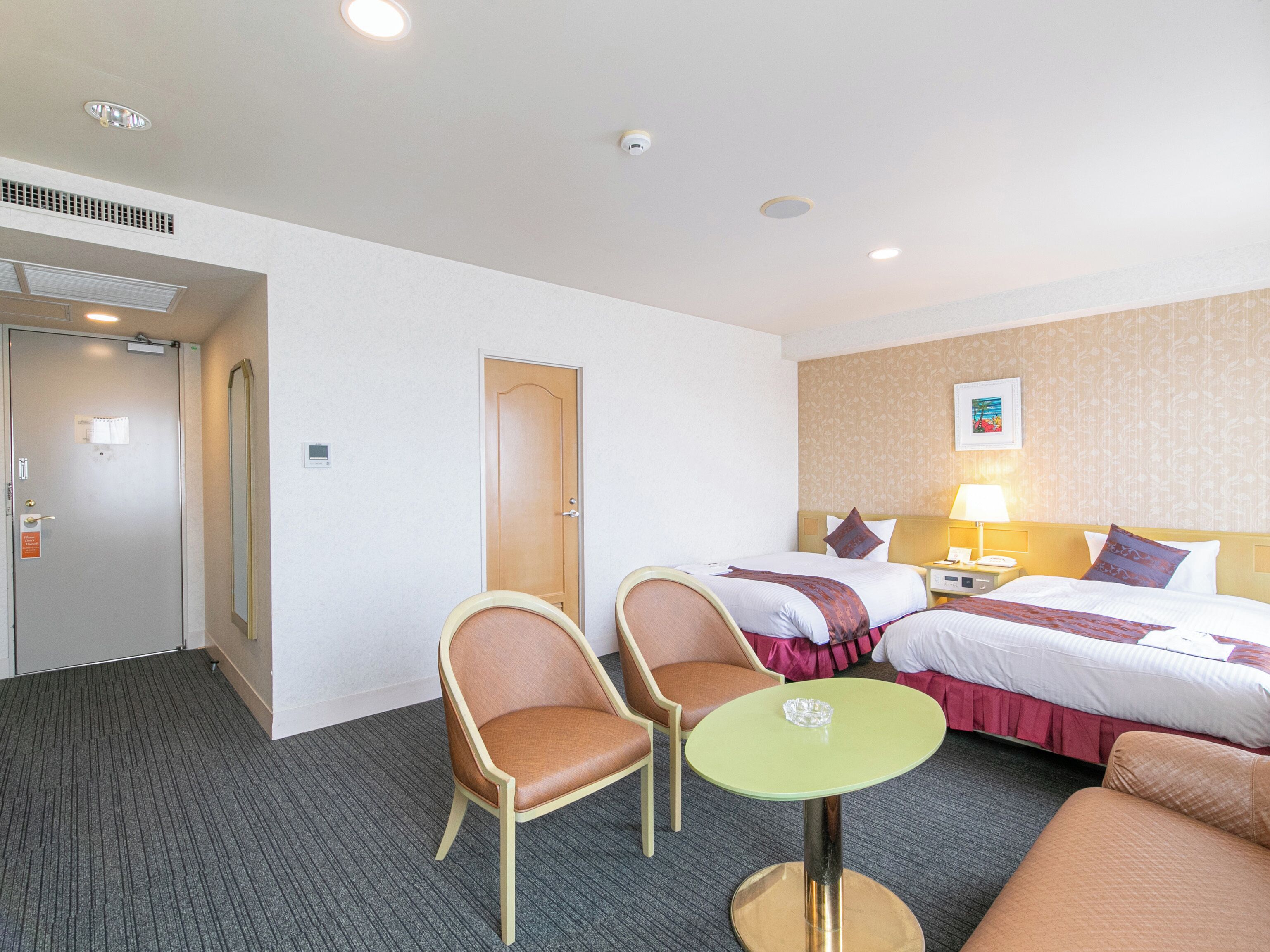 A hotel room with two single beds, a desk with a chair, a small table, and a door.