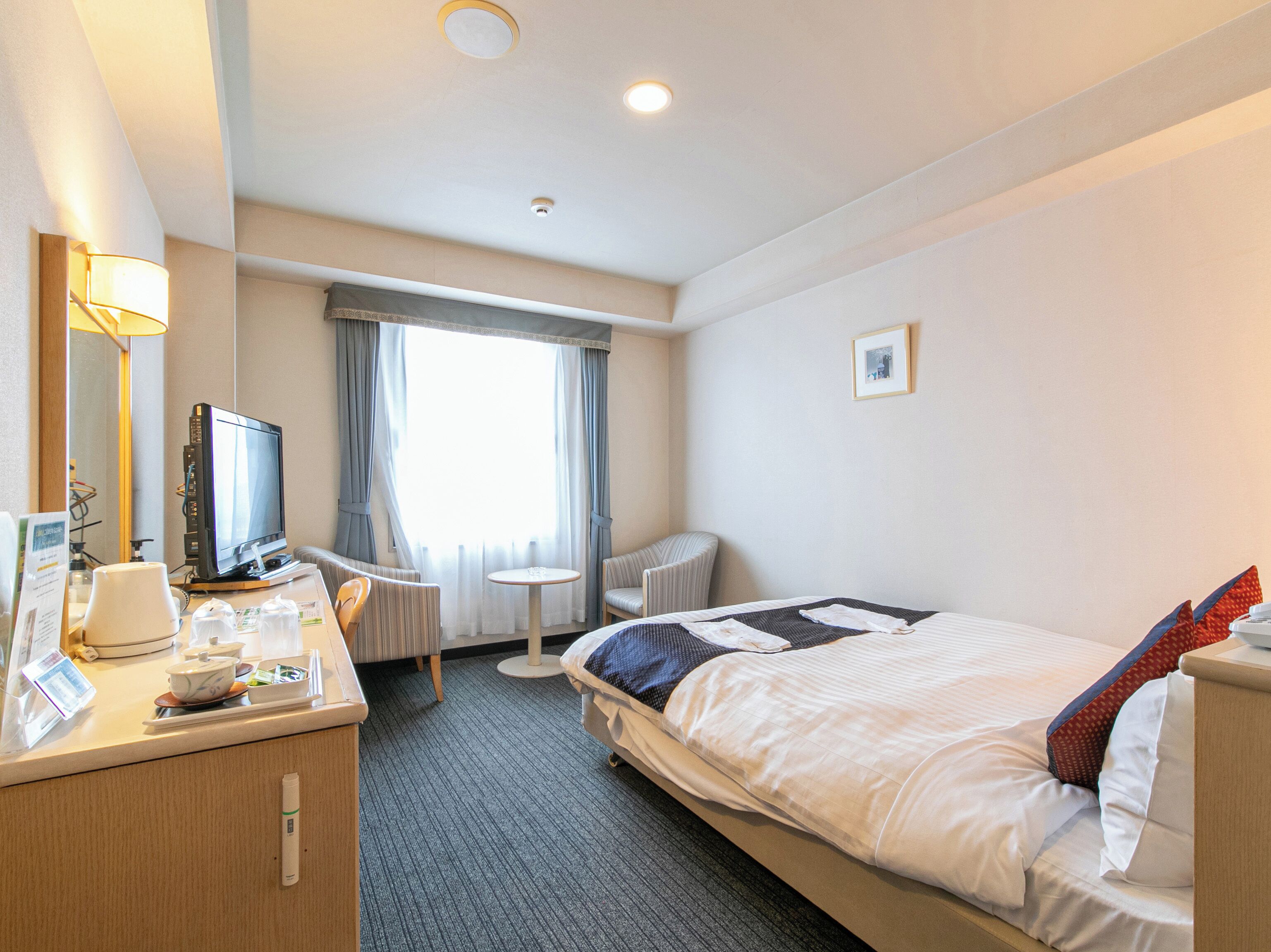 A hotel room with a bed, a desk with a computer, a chair, a small table, and a window with curtains.
