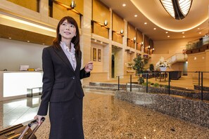 A woman in a business suit holding a suitcase in a hotel lobby with a reception desk, seating area, and a staircase. - Wakayama Urban Hotel (Wakayama)