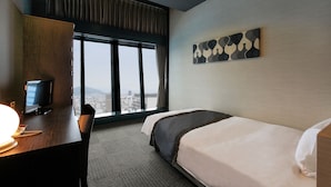Une chambre d’hôtel équipée d’un lit, d’un bureau avec un ordinateur, d’une télévision et d’une grande fenêtre donnant sur la ville.