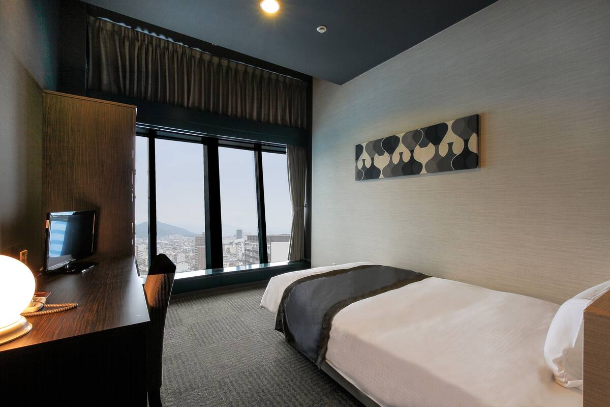 Een hotelkamer met een bed, een bureau met een computer, een televisie en een groot raam met uitzicht op de stad.