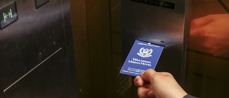 A hand holding a card with the text "Wakayama Urban Hotel" in front of an elevator with a sign above it.