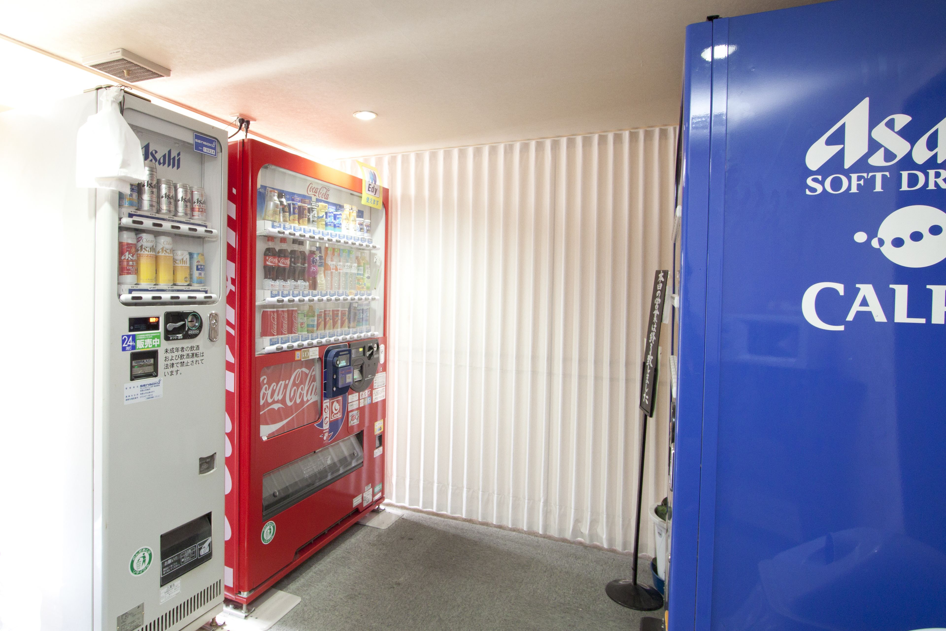 A vending machine with various drinks and snacks, a blue refrigerator with branding, and a window with vertical blinds.