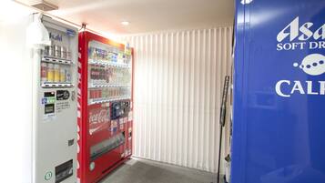 A vending machine with various drinks and snacks, a blue refrigerator with branding, and a window with vertical blinds.