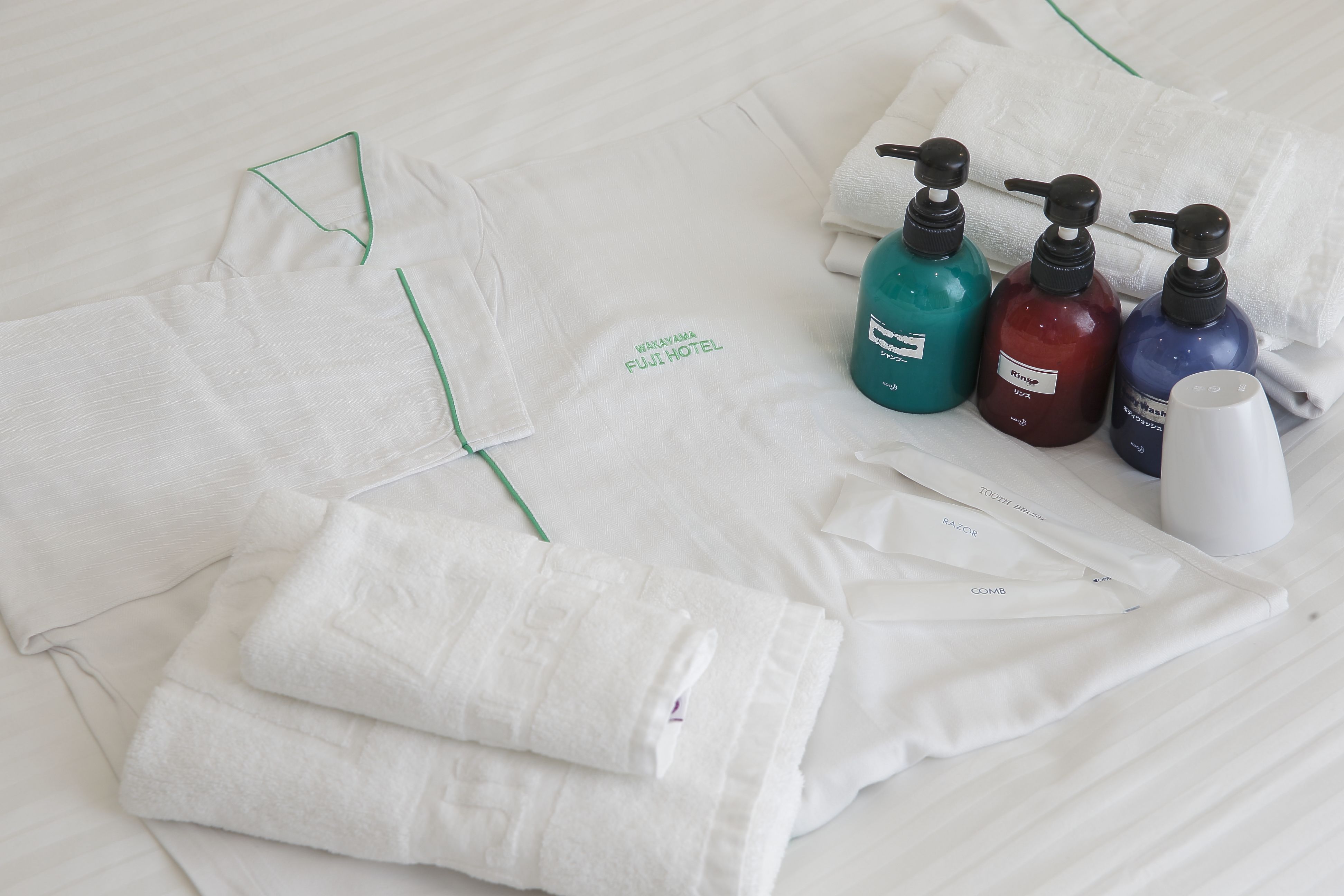 Neatly folded white towels and a white shirt with green trim on a bed. There are four bottles of liquid soap in teal, red, and blue colors.