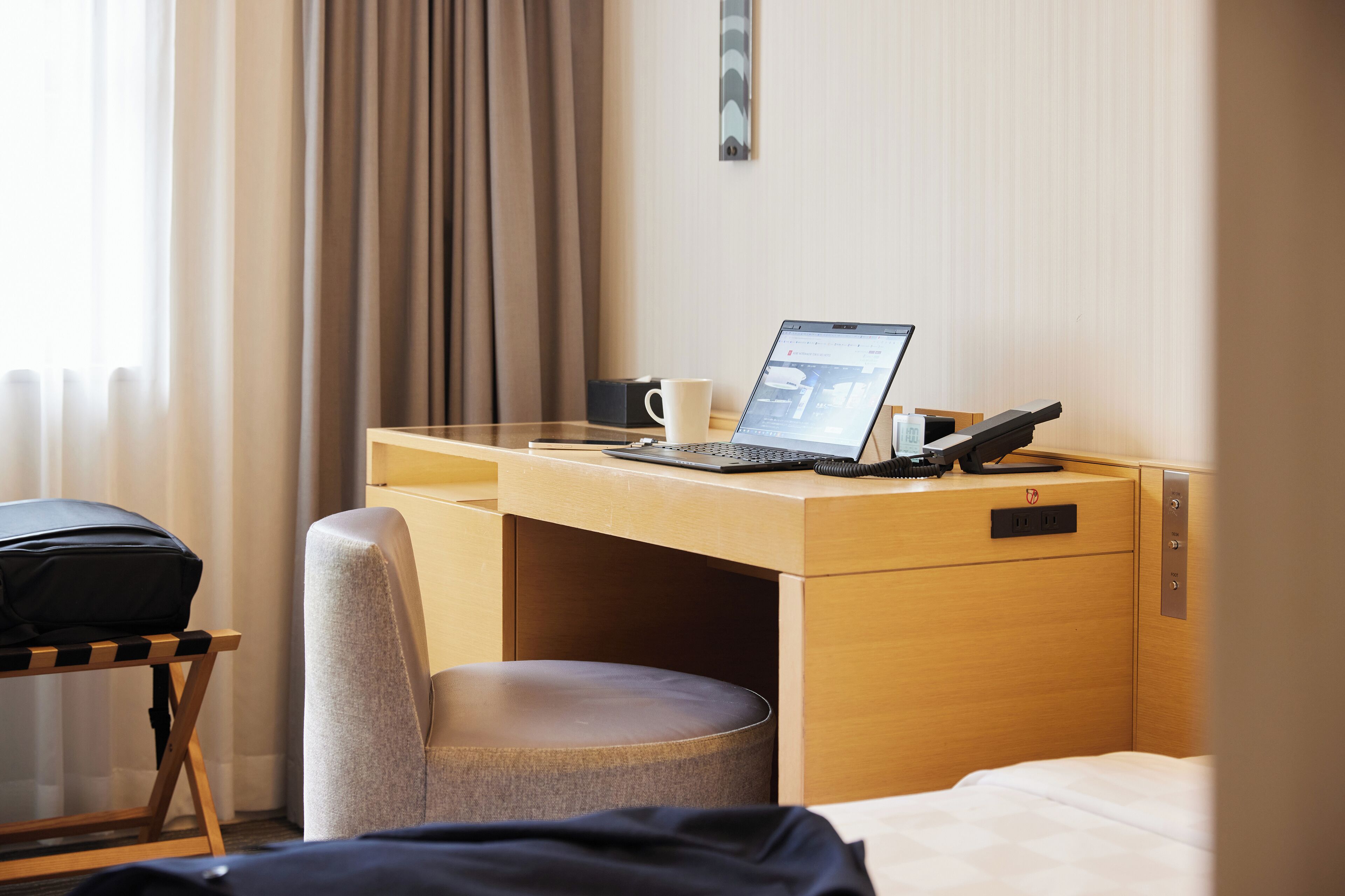 A hotel room with a wooden desk, a laptop, a telephone, and a cup.