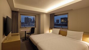 Une chambre d’hôtel avec un grand lit, un bureau, une chaise, une télévision et deux fenêtres donnant sur la ville.